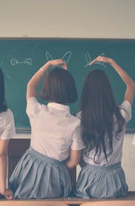 Group of teenage girls in school uniforms enjoying time together in a classroom, posing with playful gestures.