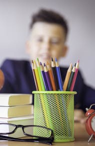 Schoolboy smiling behind a desk with books, pencils, and an alarm clock symbolizing study and creativity.