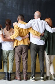 A diverse group of adults in casual outfits hugging in front of a chalkboard, symbolizing teamwork.